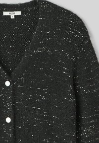 Cardigan noir en tissu tricoté texturé. Présente des paillettes éparses, un col en V, et trois boutons blancs. Étiquette de taille "S" visible.