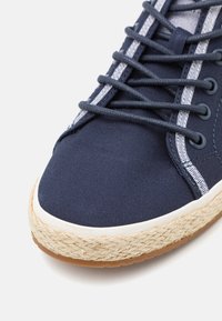Marineblå kanvas sneakers med teksturert overflate, hvit gummirand og jute-lignende såle. Har mørke snører med lyse øyelette-detaljer.