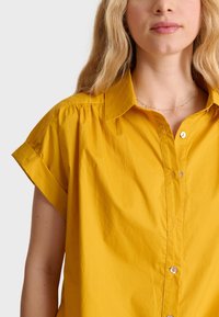 Camicia a maniche corte gialla senape con bottoni e colletto, caratterizzata da una sottile texture e una vestibilità rilassata, mostrata su uno sfondo neutro.