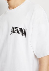 Biały bawełniany t-shirt z zaokrąglonym czarnym napisem "ACES HIGH" na piersi. Krótkie rękawy i okrągły dekolt.