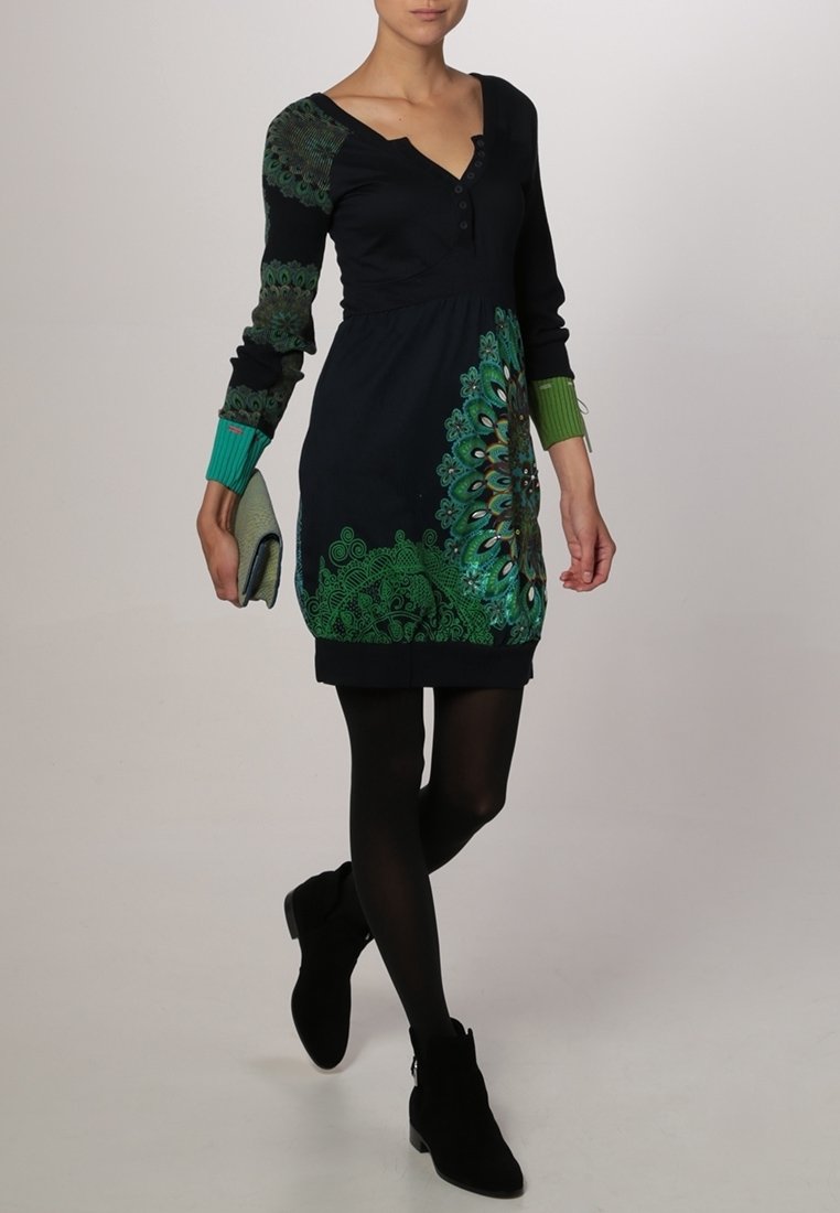 Femme portant une robe noire avec des motifs de plumes de paon verts, des collants noirs, des bottines noires et tenant une petite pochette.