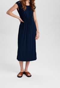 Abito blu navy fino al ginocchio con maniche a coppa, vita elastica e spacchi laterali, abbinato a sandali marroni con cinturini. Tessuto dalla texture morbida.