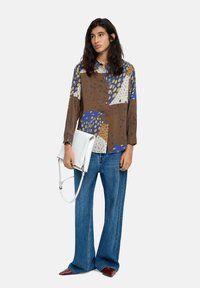 Brązowa bluzka w deseń z niebieskimi, zielonymi i szarymi akcentami, zestawiona z jasnoniebieskimi jeansami o wysokim stanie i białą torebką na ramię.