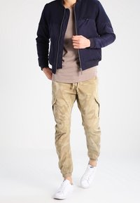 Granatowa kurtka bomberka na zamek, beżowe joggery w kamuflażu oraz białe trampki. Kurtka ma gładką teksturę i dopasowany krój.