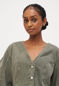 Χακί πράσινη, ελαφριά ζακέτα με V λαιμόκοψη, μεγάλα μανίκια και δύο ασημένια κουμπιά. Διαθέτει υφαντό ύφασμα και ορατές ραφές.