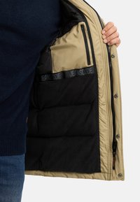 Beige jakkeinnside med svart fleecefor, glidelåslomme og en merket stripe med "JEANSCODE." Glatt, isolert materiale.