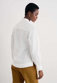 Camicia bianca a maniche lunghe con colletto alla cinese, polsini con bottone e vestibilità rilassata. Abbinata a pantaloni marroni per un contrasto.