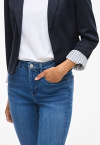 Jūrinio stiliaus džinsinis švarkas su įpjovimu ant apykaklės virš baltos marškinėlių, derinamas su šviesiai mėlynomis aukšto liemens džinsais. Švarke yra dryžiuoti rankogaliai.