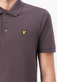 Dunkelgraues Polohemd aus strukturiertem Stoff mit einem Drei-Knopf-Verschluss, kurzen Ärmeln und einem gelben, gestickten Adler-Logo auf der Brust.