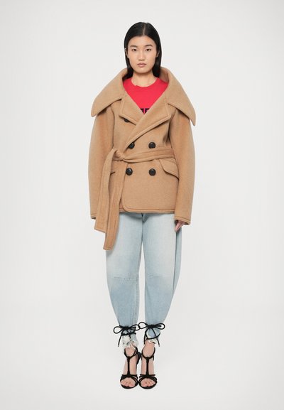 Caban en mélange de laine beige avec un large col, un devant croisé à double boutonnage et une ceinture assortie. Porté sur une chemise rouge et un jean bleu clair avec des accents de nœud.