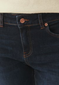 Sötétkék farmernadrágok réz gombbal és varrással, klasszikus ötlapkás kialakítással, sima textúrával és finom fakulással.