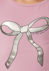 T-shirt rose avec un grand design de ruban argenté ayant un nœud central. Le texte "SUIVEZ VOS RÊVES" est à droite du ruban.