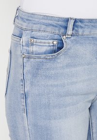 Light blue denim jeans featuring a classic five-pocket design with metal studs on the front pocket. Distressed texture and stitching details are visible.