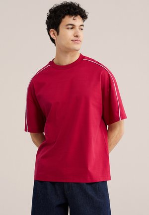 Ung mand iført rød kortærmet skjorte med hvide striber på skuldrene, kigger sidelæns, stående mod en ensfarvet baggrund.