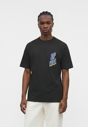 Νέος άνδρας με πλεκτά μαλλιά που φοράει μαύρο T-shirt με μπλε και κίτρινο κείμενο "RIP CURL" και λευκό παντελόνι, στέκεται μπροστά σε απλό λευκό φόντο.