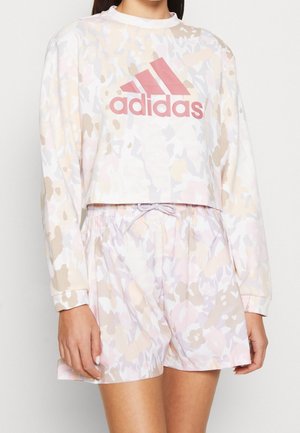 Kobieta nosząca pastelową długorękawową krótka bluzę w kamuflażowy wzór z różowym logo Adidas oraz pasujące krótkie spodenki ze ściągaczem w talii.