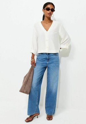 Femme portant un chemisier blanc boutonné, un jean bleu à jambes larges, des sandales à talons marron, des lunettes de soleil, des boucles d’oreilles créoles, tenant un sac en daim marron.