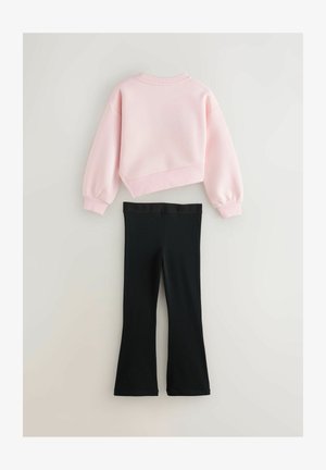 Rozā kroptā sporta jaka ar garām piedurknēm, pārī ar melnām elastīgām legingām ar melnu elastīgu jostiņu, kurā ir uzraksts "BAKE".