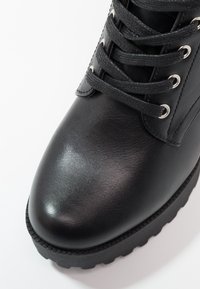 Botín de cuero negro hasta el tobillo con acabado texturizado, puntera redonda y suela de goma gruesa. Presenta un diseño de cordones y ojales metálicos.