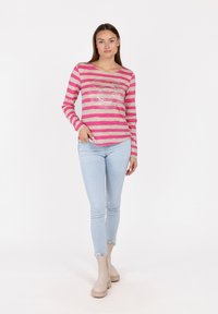 Langarmoberteil mit pink- und beigen Streifen, runder Halsauschnitt und einem metallischen Grafikdruck. Kombiniert mit hellblauen Jeans und beigen Stiefeln.