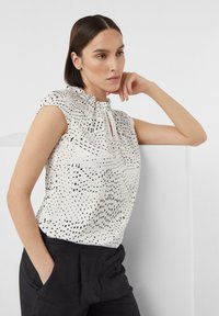 White blouse with black dot pattern, short sleeves, gathered neckline, and keyhole detail at the front. Worn with black trousers.