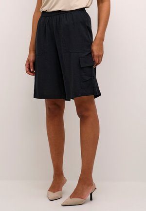 Kvinde iført sorte cargo shorts til knæet med elastisk talje og beige højhælede sko, stående mod en ensfarvet baggrund.