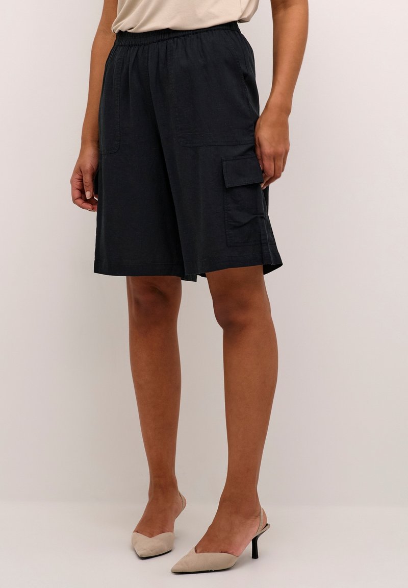 Femme portant un short cargo noir longueur genou avec une taille élastique et des chaussures à talons beiges, debout devant un fond uni.