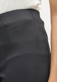 Personne portant un pantalon noir taille haute et ajusté et un haut blanc, montrée de la taille au haut de la cuisse, debout contre un fond uni.