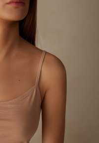 Beige těsně padnoucí tílko s tenkými ramínky, hladkým povrchem a standardním kulatým výstřihem, zobrazené na jemném, neutrálním pozadí.