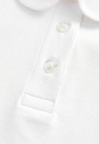 Valge polo särk, millel on tekstuuriga kangas, klassikaline krae ja kaks südamekujulist selget nõela koos logo detailiga.