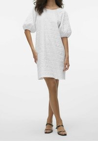 Valkoinen lyhyt mekko, jossa on teksturoitu kangas, pyöreä pääntie ja puffi hihat. Yhdistetty mustiin hihnasaappaisiin.