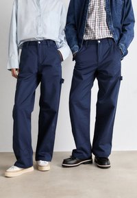 Twee modellen dragen marineblauwe katoenen broeken met een losse pasvorm, zijzakken en contrasterende schoenen - de ene beige en de andere zwart.