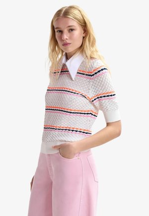 Giovane donna con capelli biondi che indossa un maglione bianco testurizzato con strisce colorate e un grande colletto bianco, abbinato a pantaloni rosa chiaro.