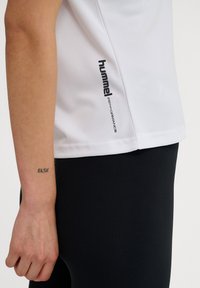 Vit t-shirt med korta ärmar i ett mjukt tyg, med en svart "hummel"-logotyp och texten "performance" på sidan. Svarta leggings synliga.