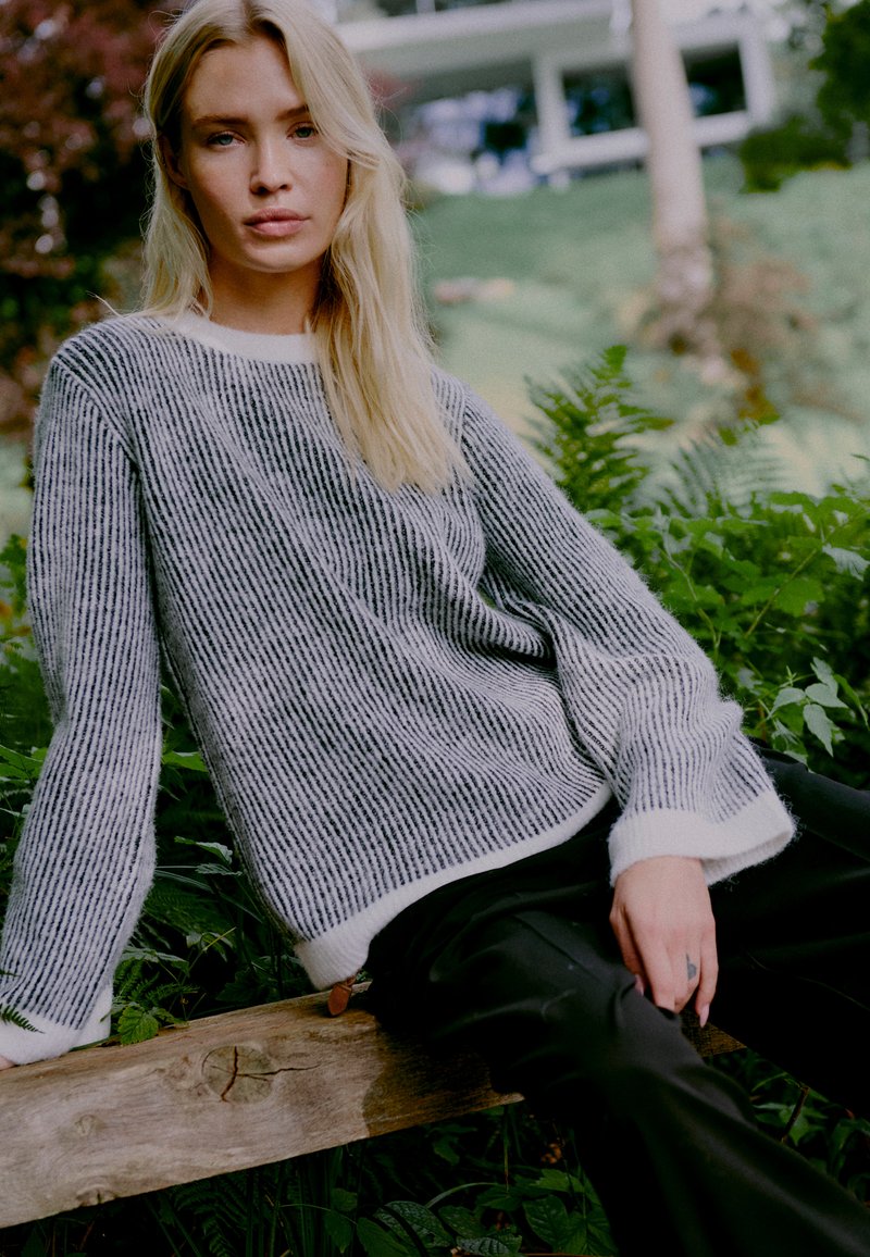 Hallide ja valgete triipudega kudum, millel on vabalt langetatud lõige ja laiad varrukad, kombineerituna mustade pükstega, rohelise ja lehtede täis väliskeskkonna taustal.