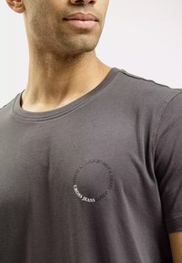 T-shirt w kolorze węglowym szarym z mieszanki bawełny, z okrągłym dekoltem. Posiada okrągłą grafikę z tekstem wokół centrum oraz nazwą marki na dole.
