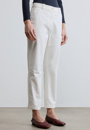 Kobieta ubrana w białe spodnie o prostych nogawkach, granatowy długi rękaw i bordowe płaskie buty, stojąca na szarej podłodze na tle jednolitej ściany.