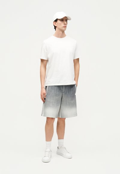 Νεαρός άνδρας που στέκεται, φορώντας λευκό καπέλο, απλό λευκό μπλουζάκι, σορτς με γκρι διαβάθμιση, λευκές κάλτσες και λευκά αθλητικά παπούτσια πάνω σε ανοιχτό φόντο.