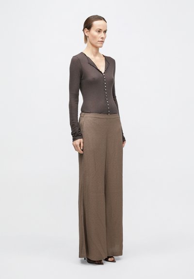 Donna in piedi, di fronte, che indossa una maglia marrone scuro con bottoni e maniche lunghe, pantaloni ampi color marrone chiaro e scarpe con tacco scure e punta aperta.