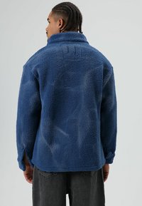 Blauwe fleecejas met een gestructureerde afwerking, hoge kraag en zijschuiven. De achterkant heeft gestikte accenten en een losse pasvorm.