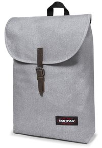 Szary materiałowy plecak z klapą na zawiasach, brązowym skórzanym paskiem z klamrą oraz czarnym logo Eastpak. Prostokątny kształt o gładkiej teksturze.