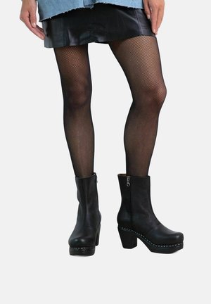 Schwarze Leder Stiefeletten mit einem robusten Absatz, strukturiertem Finish, seitlichem Reißverschluss und dekorativer Naht entlang der Sohle. Kombiniert mit Netzstrumpfhosen.