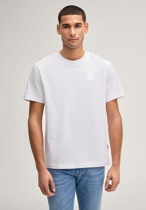 Junger Mann mit kurzem Haar, der ein schlichtes weißes T-Shirt mit kurzen Ärmeln und blaue Jeans trägt, vor hellem neutralem Hintergrund stehend.