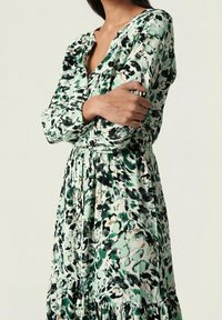 Vestido de estampado floral en tonos de verde, negro y crema; mangas largas, escote en forma de V, cintura ceñida y falda en capas. Textura de tela suave.