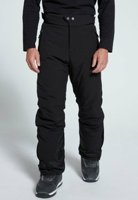 Schwarze isolierte Hose mit hohem Bund, Druckknopfverschluss, seitlichen Reißverschlusstaschen und leicht tailliertem Bein. Getragen mit grauen Stiefeln.