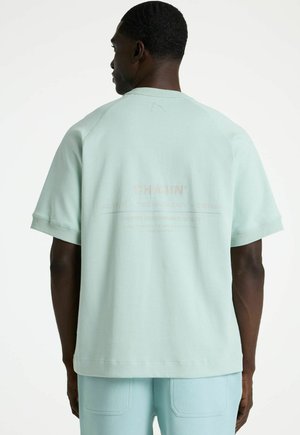 Man met kort haar die een lichtblauw oversized shirt met korte mouwen en bijpassende broek draagt, met de rug naar de camera tegen een witte achtergrond.