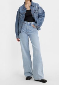 Personne portant une veste en jean bleue ample, un débardeur noir, un jean large bleu clair et des chaussures noires à bout carré, debout avec une main dans la poche.