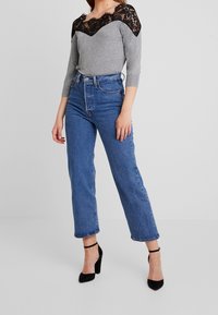 Haut gris à manches longues avec encolure en dentelle noire, associé à un jean bleu taille haute et des escarpins à brides noires.