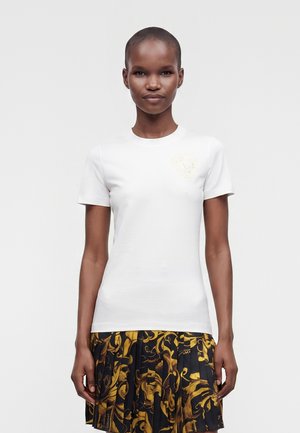 Ung kvinde med kort hår iført en hvid tætsiddende T-shirt og et plisseret sort skørt med gule blomsterprint, stående mod en ensfarvet baggrund.