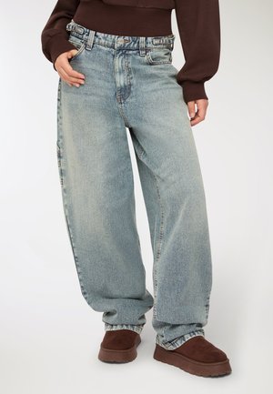 Persona che indossa jeans larghi blu chiaro con fibbie laterali regolabili, un maglione corto marrone e scarpe in suede marroni in piedi.